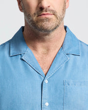 A close-up shows a man wearing the Psycho Bunny MENS BIG AND TALL KERVIN LIGHTWEIGHT DENIM WOVEN SHIRT (B9Q467F200), with only his lower face and upper torso in frame for a casual look.