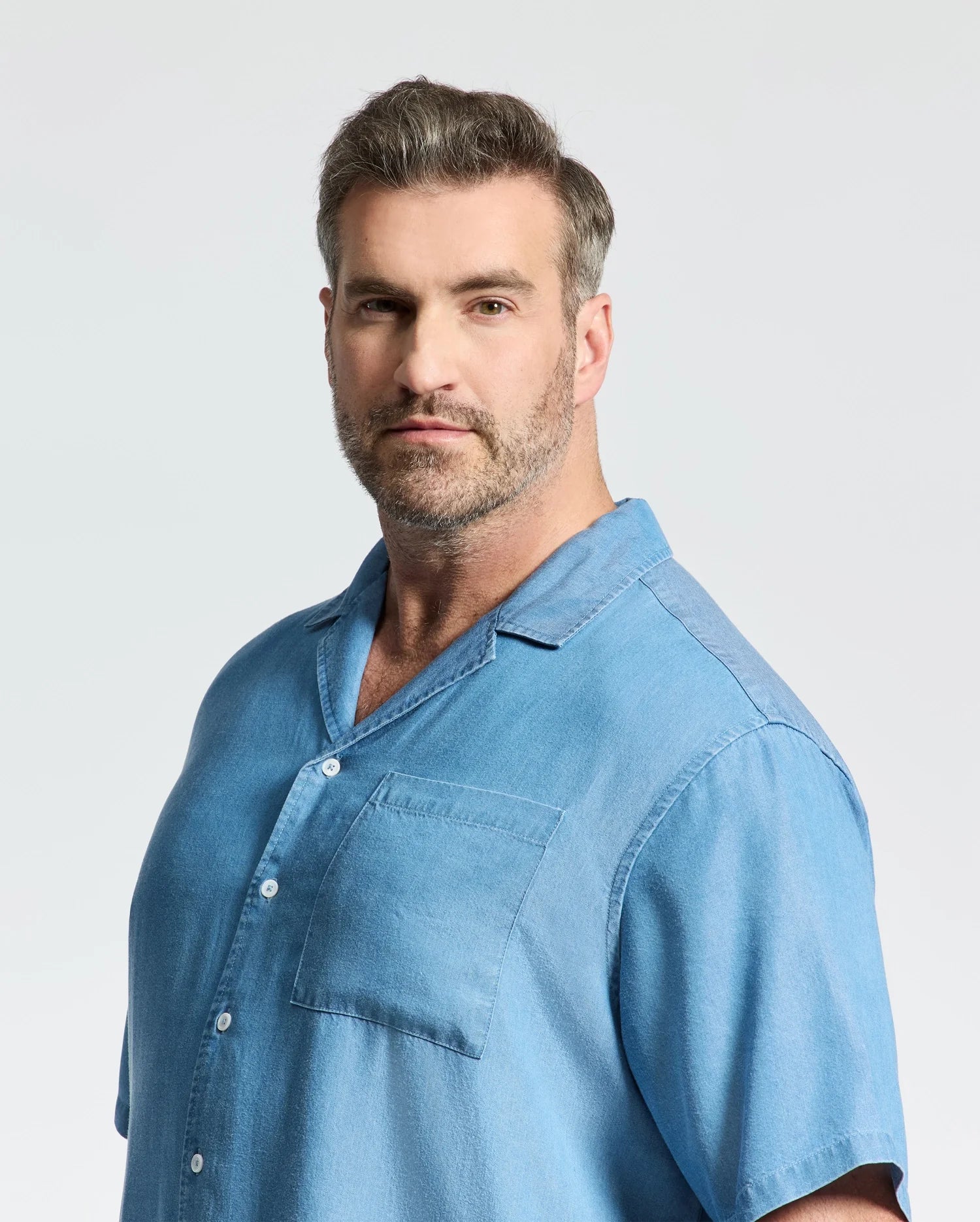A man with short gray hair and a trimmed beard models the Psycho Bunny MENS BIG AND TALL KERVIN LIGHTWEIGHT DENIM WOVEN SHIRT (B9Q467F200), standing casually against a plain, light background.