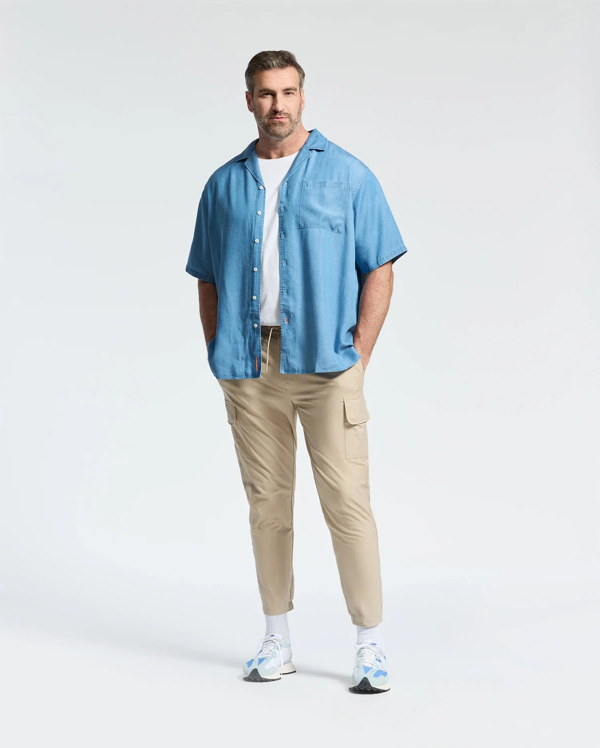 A man with short hair and a beard stands hands in pockets, wearing the Psycho Bunny MENS BIG AND TALL KERVIN LIGHTWEIGHT DENIM WOVEN SHIRT (B9Q467F200) over a white tee, with beige cargo pants and white sneakers for a relaxed, casual style.