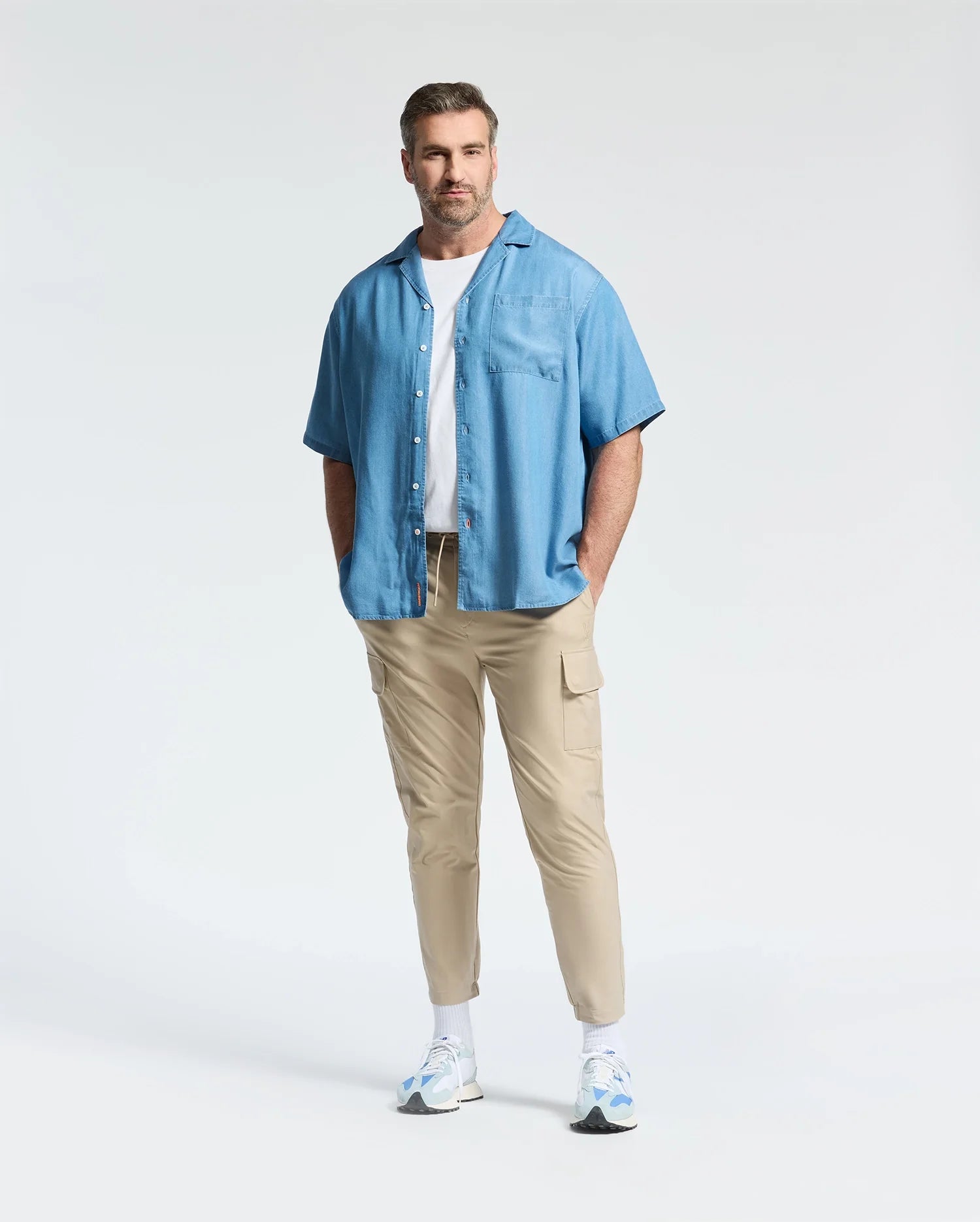 A man with short hair and a beard stands hands in pockets, wearing the Psycho Bunny MENS BIG AND TALL KERVIN LIGHTWEIGHT DENIM WOVEN SHIRT (B9Q467F200) over a white tee, with beige cargo pants and white sneakers for a relaxed, casual style.