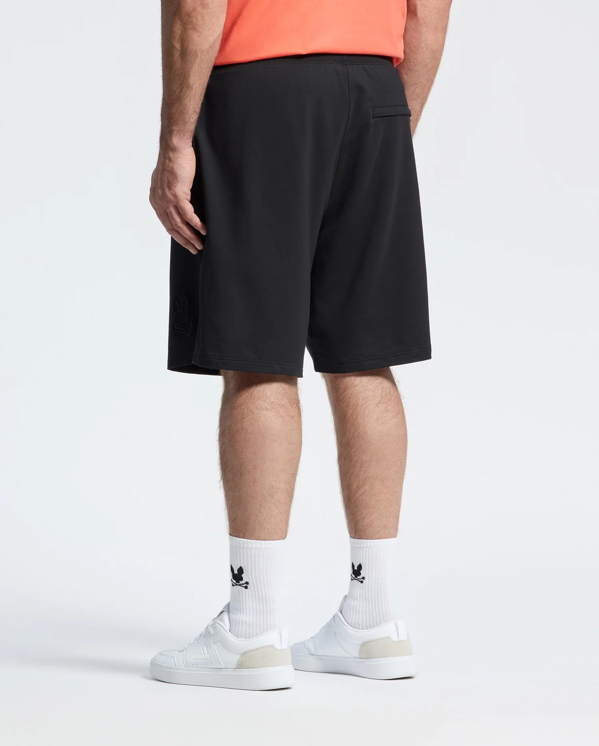 A person stands against a plain white background, seen from the back, wearing a coral shirt, white sneakers, white socks with black Adidas logos, and Psycho Bunny MENS BIG AND TALL LEVANZO EMBROIDERED MICRO FRENCH TERRY SWEATSHORTS.