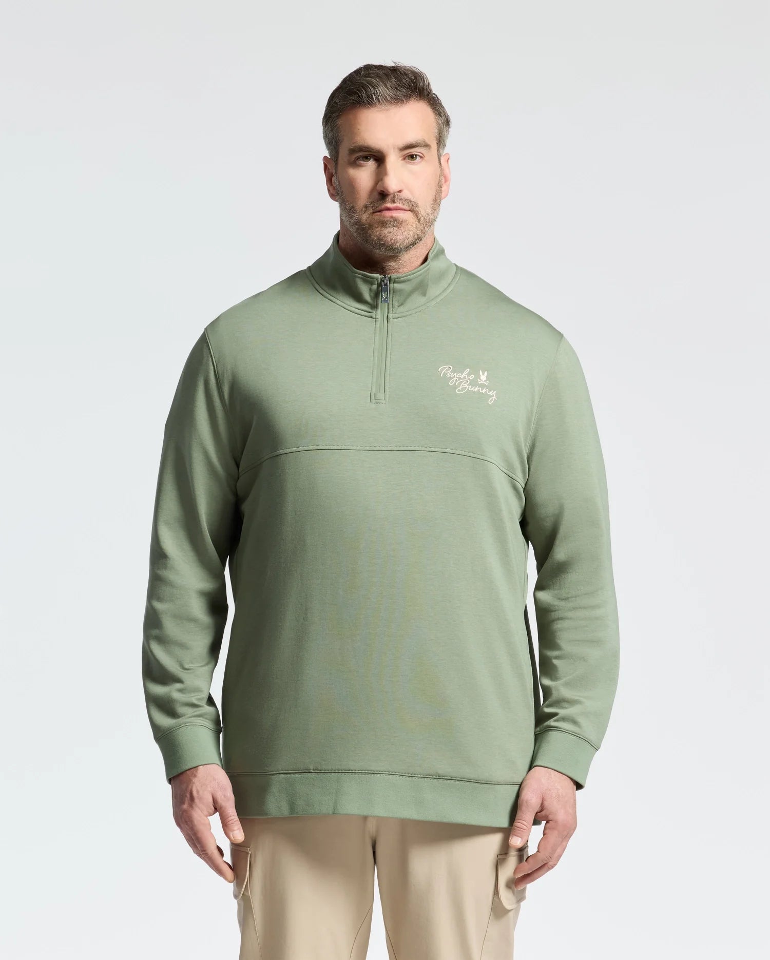 A man stands against a white background wearing the Psycho Bunny “Mens Big and Tall Desert 1/4 Mock Zip - B9S488F200,” a green cotton quarter-zip pullover with paneled construction and an embroidered logo. His short hair complements his light-colored pants, completing the stylish ensemble.