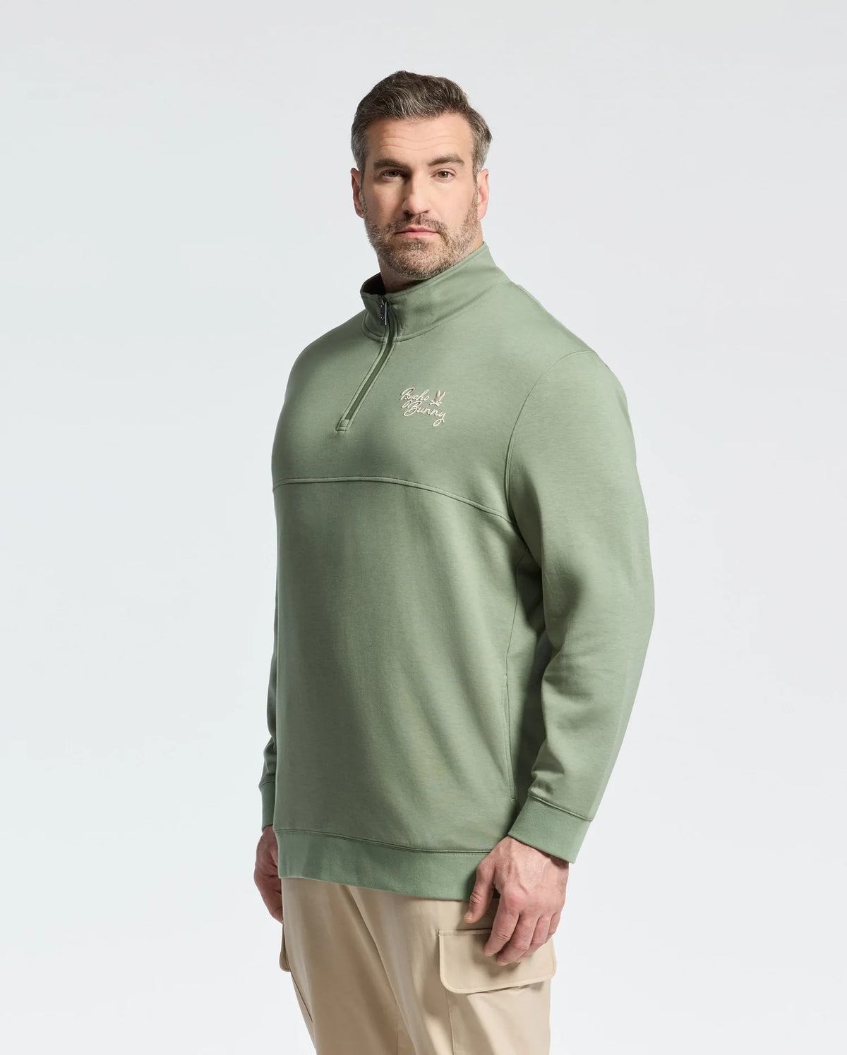A man with short hair and a beard faces the camera confidently, wearing Psycho Bunny's green cotton MENS BIG AND TALL DESERT 1/4 MOCK ZIP - B9S488F200, featuring an embroidered chest logo, paired with khaki pants against a plain backdrop.