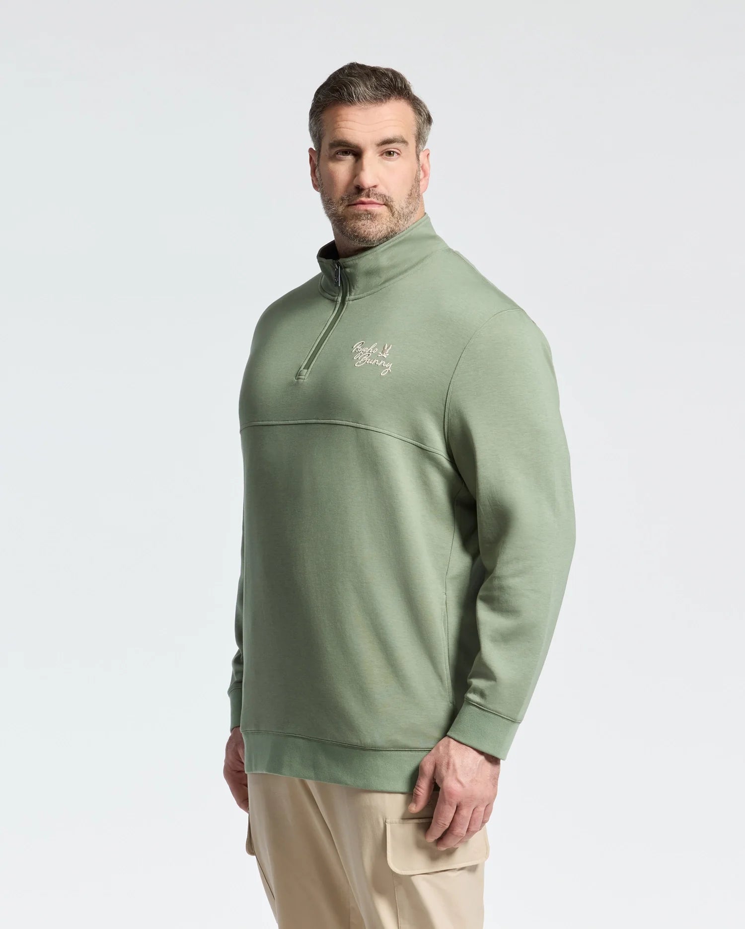 A man with short hair and a beard faces the camera confidently, wearing Psycho Bunny's green cotton MENS BIG AND TALL DESERT 1/4 MOCK ZIP - B9S488F200, featuring an embroidered chest logo, paired with khaki pants against a plain backdrop.