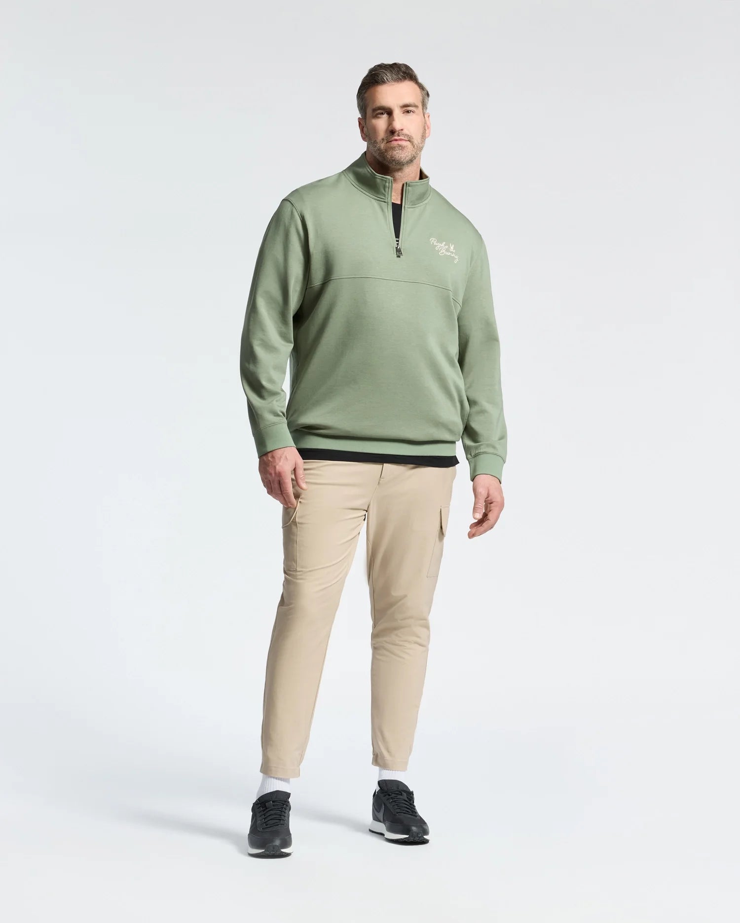 A man stands against a light gray background, wearing a Psycho Bunny green cotton quarter-zip sweatshirt (MENS BIG AND TALL DESERT 1/4 MOCK ZIP - B9S488F200) with paneled construction and an embroidered chest logo, beige pants, and black sneakers with white soles. He has short hair and a beard.