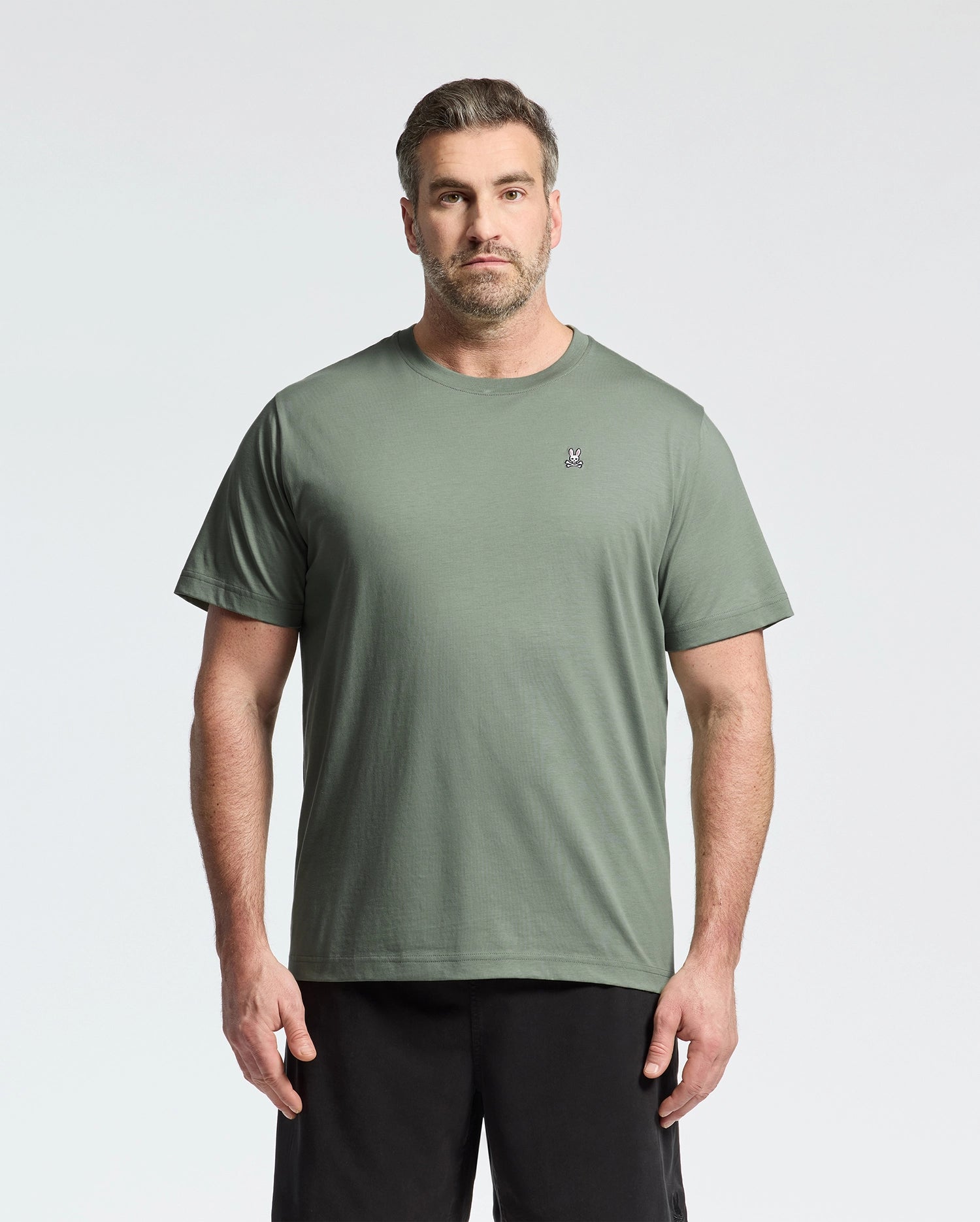 A man wears a sage green BIG AND TALL CLASSIC CREW NECK TEE from Psycho Bunny, made of 100% Pima cotton and featuring a small embroidered chest logo. He pairs it with black shorts, standing against a white background, sporting short hair and a beard.