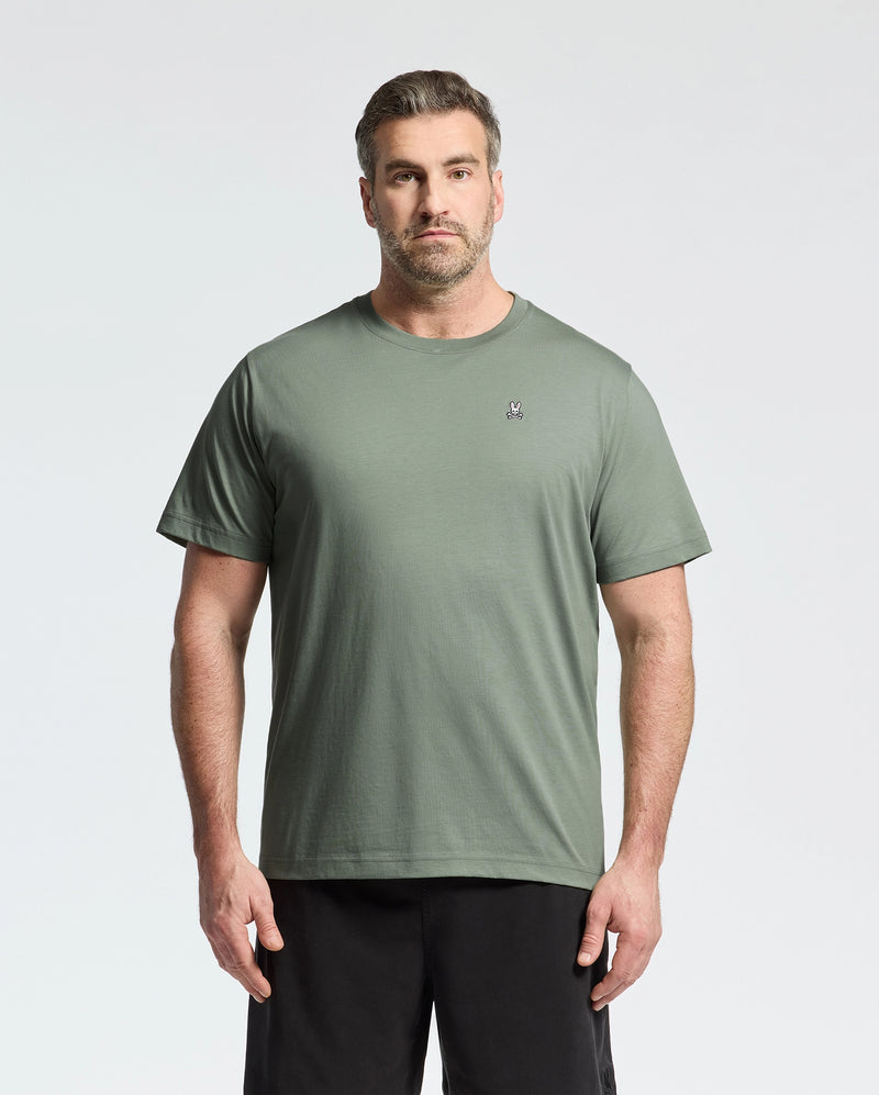 A man wears a sage green BIG AND TALL CLASSIC CREW NECK TEE from Psycho Bunny, made of 100% Pima cotton and featuring a small embroidered chest logo. He pairs it with black shorts, standing against a white background, sporting short hair and a beard.