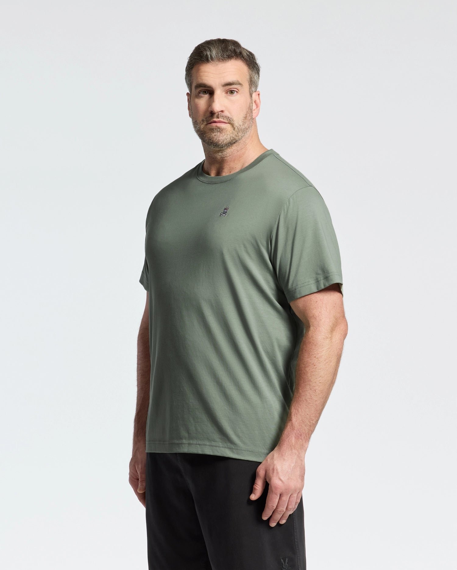 A man with short hair and a beard wears a Psycho Bunny BIG AND TALL CLASSIC CREW NECK TEE in olive green, made from 100% Pima cotton, paired with black pants; he stands against a plain white background.