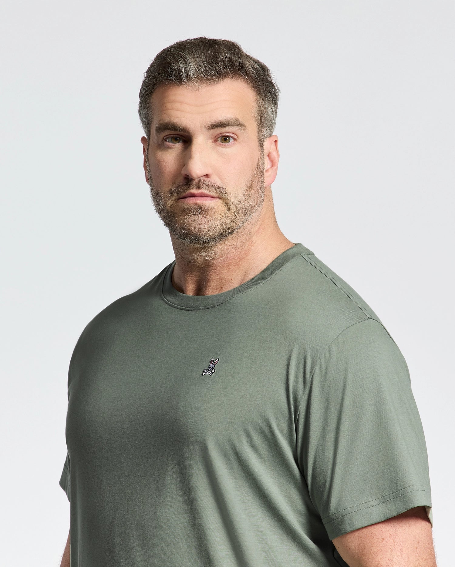 A bearded man with short brown hair wears the Psycho Bunny BIG AND TALL CLASSIC CREW NECK TEE - B9U014CRPC in green, made from 100% Pima cotton, featuring a small chest embroidery. He faces the camera against a light backdrop.