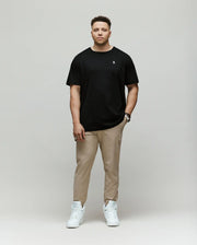 A man faces forward against a plain background, wearing the Psycho Bunny MENS BIG AND TALL CLASSIC CREW NECK TEE - B9U014CRPC in black, paired with beige pants and white sneakers. His left hand is in his pocket and he has a neutral expression.