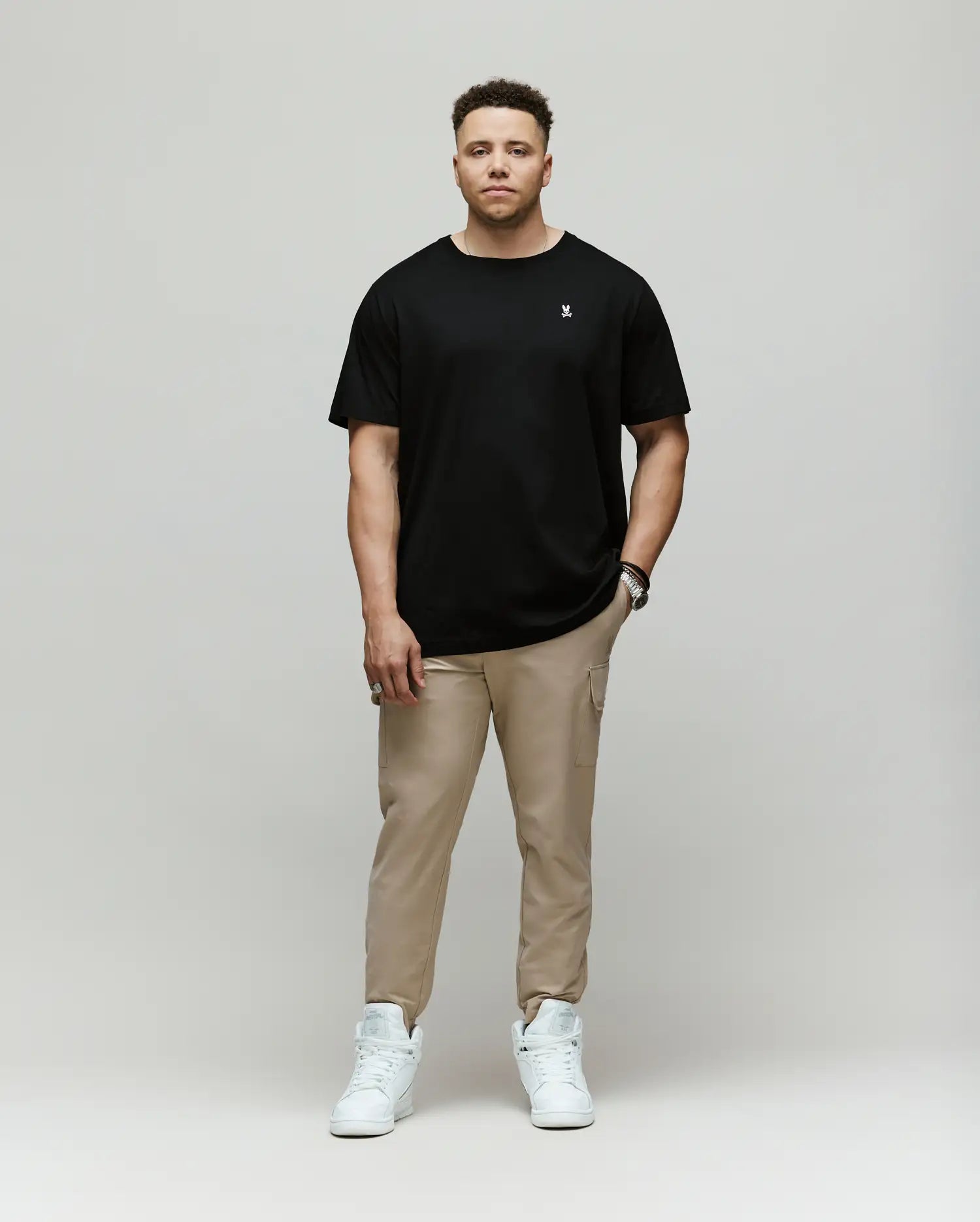 A man faces forward against a plain background, wearing the Psycho Bunny MENS BIG AND TALL CLASSIC CREW NECK TEE - B9U014CRPC in black, paired with beige pants and white sneakers. His left hand is in his pocket and he has a neutral expression.
