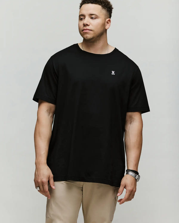 A man with short curly hair wears beige pants and a Psycho Bunny MENS BIG AND TALL CLASSIC CREW NECK TEE - B9U014CRPC, a loose black tee with a small embroidered logo on the left chest, against a plain background. He has a watch on his left wrist.