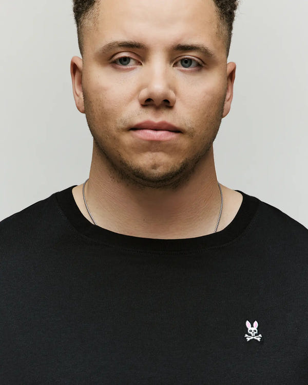 A man with light brown skin and short curly hair wears the Psycho Bunny Men's Big and Tall Classic Crew Neck Tee (B9U014CRPC), 100% Pima cotton, with a small bunny and crossbones pin on the chest, standing against a plain background.