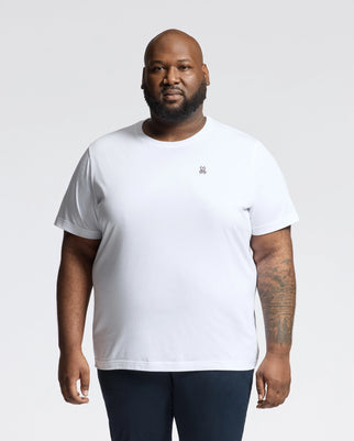 A bearded man wears the Psycho Bunny Men's Big and Tall Classic Crew Neck Tee in pure white, featuring a small embroidered logo on the left. Made from soft Pima cotton, this tee pops against the light gray backdrop.