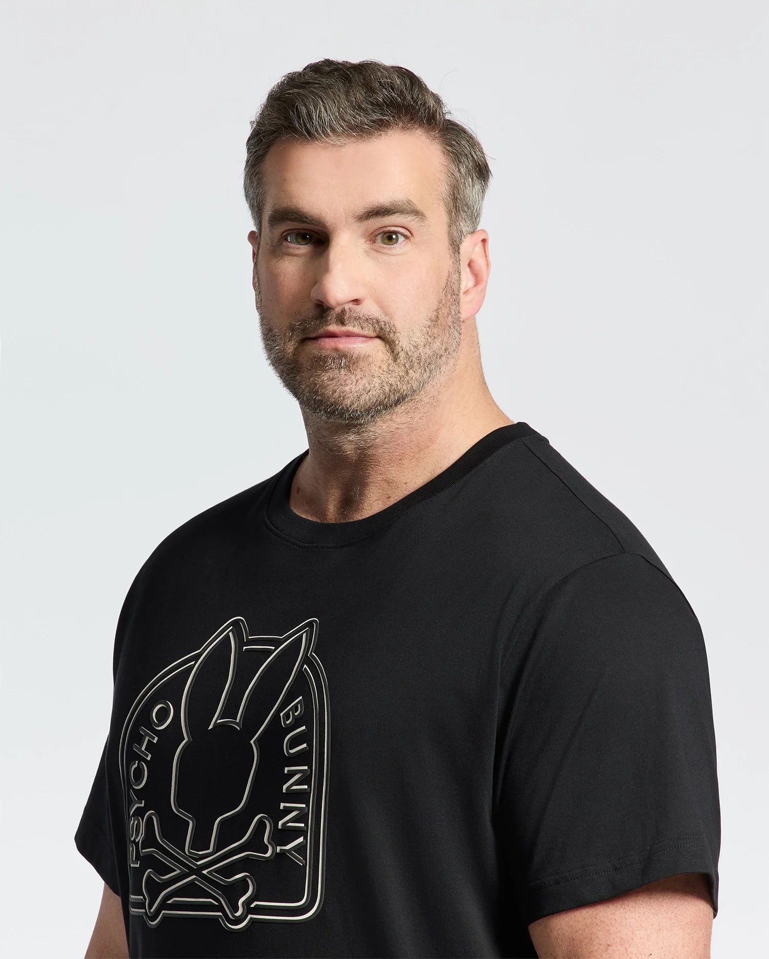 A man with short hair and a beard wears the Psycho Bunny Men's Big and Tall Melbourne Graphic Tee, crafted from eco-friendly Peruvian Pima cotton. The black T-shirt showcases a bunny with crossbones and "PSYCHO BUNNY" text. The background is plain and light-colored.
