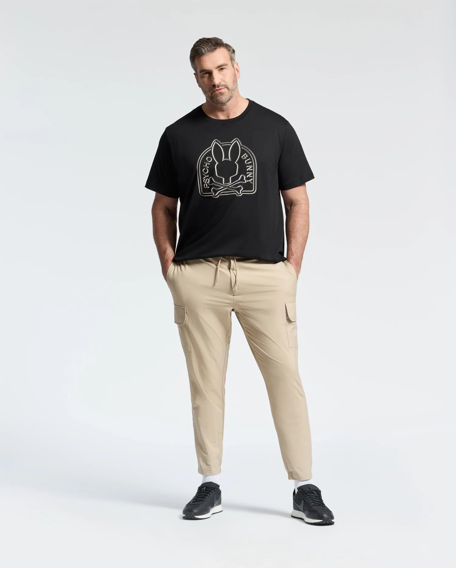 A man stands against a white background wearing Psycho Bunny's eco-friendly black "Melbourne Graphic Tee" in soft Peruvian Pima cotton featuring a bunny graphic, paired with beige cargo pants and black sneakers, hands in pockets.