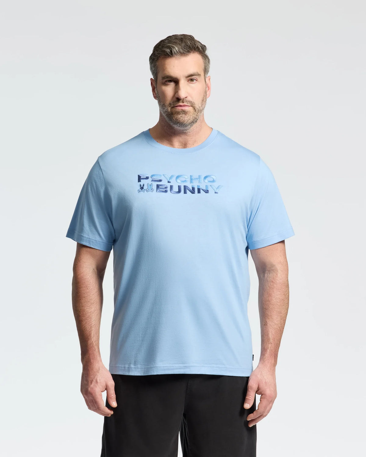 A man with short hair and a beard wears the Psycho Bunny MENS BIG AND TALL BERNI EMBROIDERED WORDMARK GRAPHIC TEE in light blue and black shorts, standing against a plain light background with a neutral expression.