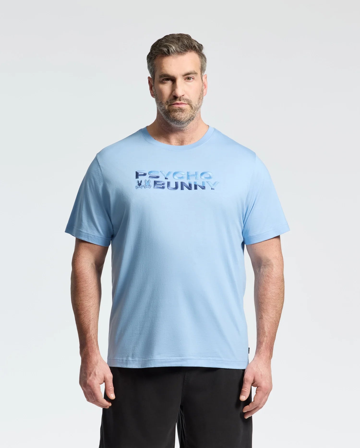 A man with short hair and a beard wears the Psycho Bunny MENS BIG AND TALL BERNI EMBROIDERED WORDMARK GRAPHIC TEE in light blue and black shorts, standing against a plain light background with a neutral expression.