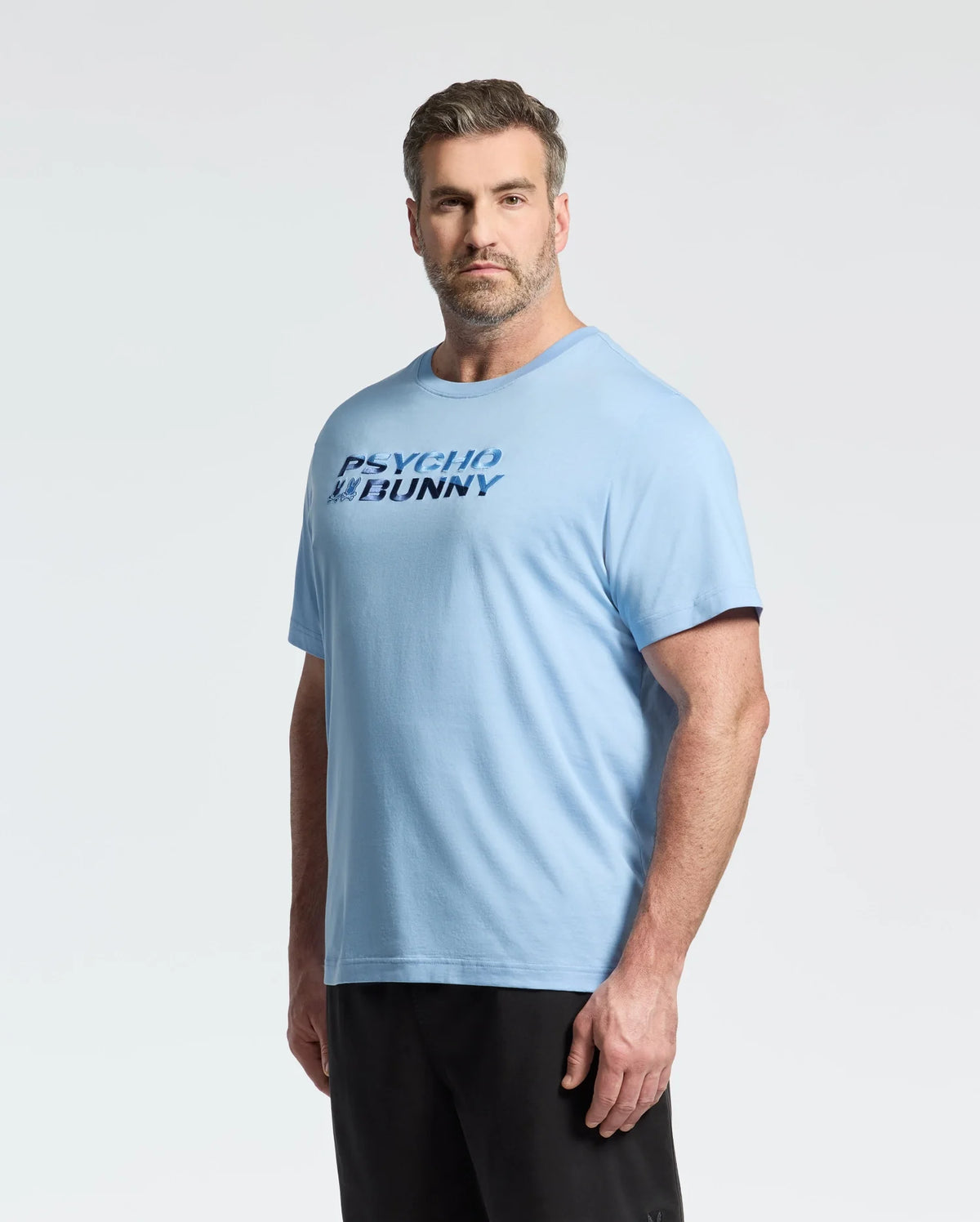 A man with short hair and a beard wears the Psycho Bunny Mens Big and Tall Berni Embroidered Wordmark Graphic Tee (B9U417F200), a light blue, breathable Pima cotton shirt with bold "PSYCHO BUNNY" lettering on the front, against a plain background.