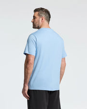 A man with short brown hair and a beard stands facing away from the camera, wearing the Psycho Bunny MENS BIG AND TALL BERNI EMBROIDERED WORDMARK GRAPHIC TEE - B9U417F200 in light blue and black shorts against a plain, light background.