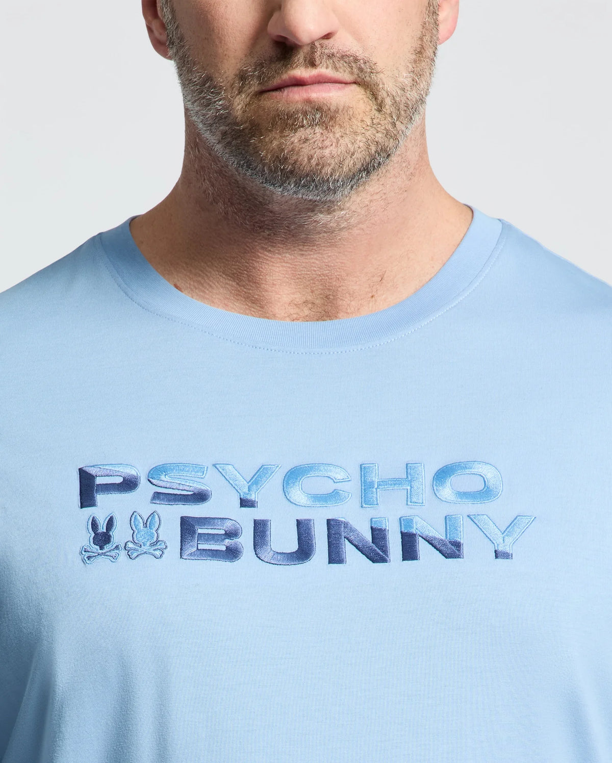 Close-up of a man in the Psycho Bunny MEN'S BIG AND TALL BERNI EMBROIDERED WORDMARK GRAPHIC TEE (B9U417F200), a light blue breathable Pima cotton tee with "PSYCHO BUNNY" and two small embroidered bunny logos on the chest.