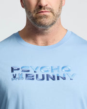 Close-up of a man in the Psycho Bunny MEN'S BIG AND TALL BERNI EMBROIDERED WORDMARK GRAPHIC TEE (B9U417F200), a light blue breathable Pima cotton tee with "PSYCHO BUNNY" and two small embroidered bunny logos on the chest.