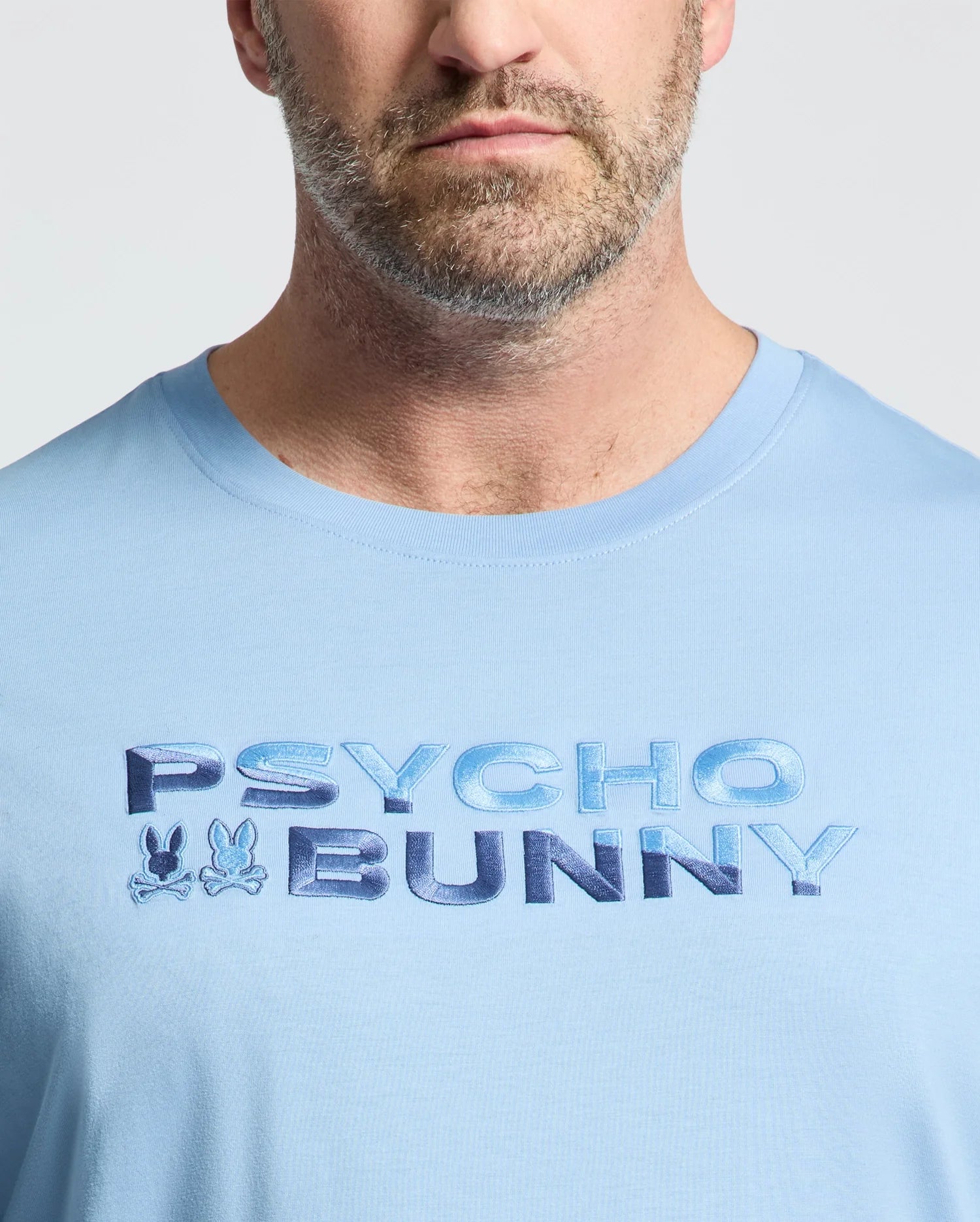 Close-up of a man in the Psycho Bunny MEN'S BIG AND TALL BERNI EMBROIDERED WORDMARK GRAPHIC TEE (B9U417F200), a light blue breathable Pima cotton tee with "PSYCHO BUNNY" and two small embroidered bunny logos on the chest.