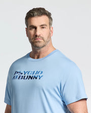 A man with short gray hair and a beard wears the Psycho Bunny Mens Big and Tall Berni Embroidered Wordmark Graphic Tee (B9U417F200) in light blue, standing against a plain, light background.