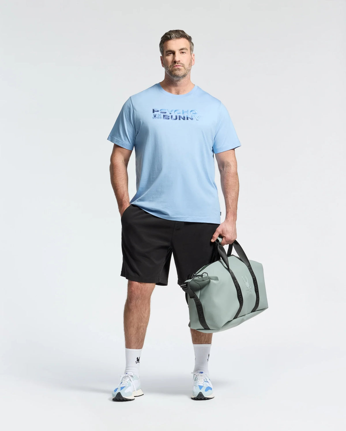 A man wears the Psycho Bunny MENS BIG AND TALL BERNI EMBROIDERED WORDMARK GRAPHIC TEE, black shorts, and white sneakers, holding a light gray duffel bag against a plain light background.