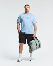 A man wears the Psycho Bunny MENS BIG AND TALL BERNI EMBROIDERED WORDMARK GRAPHIC TEE, black shorts, and white sneakers, holding a light gray duffel bag against a plain light background.