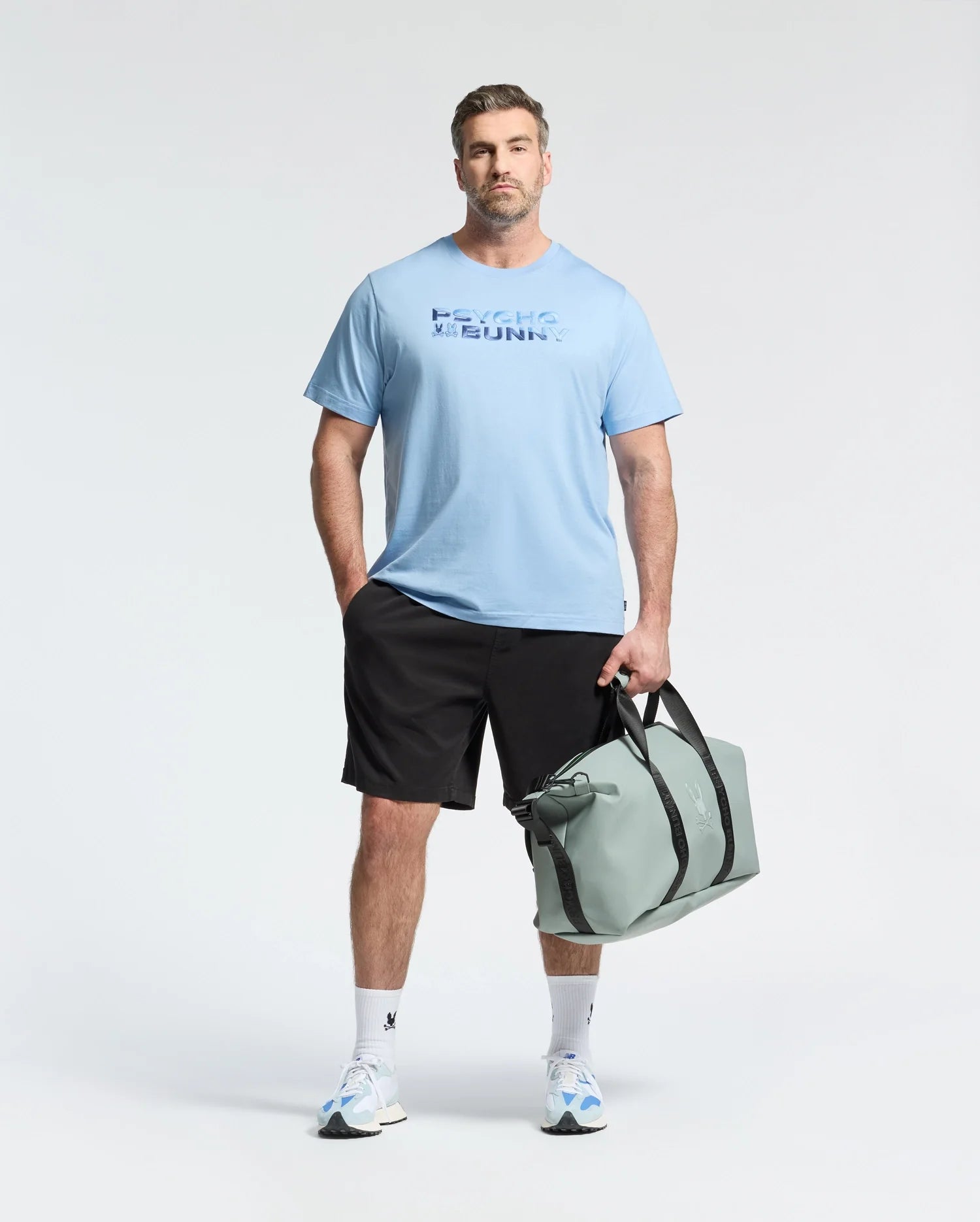 A man wears the Psycho Bunny MENS BIG AND TALL BERNI EMBROIDERED WORDMARK GRAPHIC TEE, black shorts, and white sneakers, holding a light gray duffel bag against a plain light background.