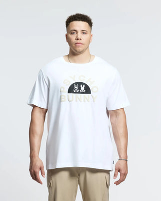 A man stands against a plain background wearing the Psycho Bunny Men's Big and Tall Goldie Graphic Tee in crisp white Pima cotton, featuring "PSYCHO BUNNY" text and a playful bunny graphic, paired with stylish beige pants.
