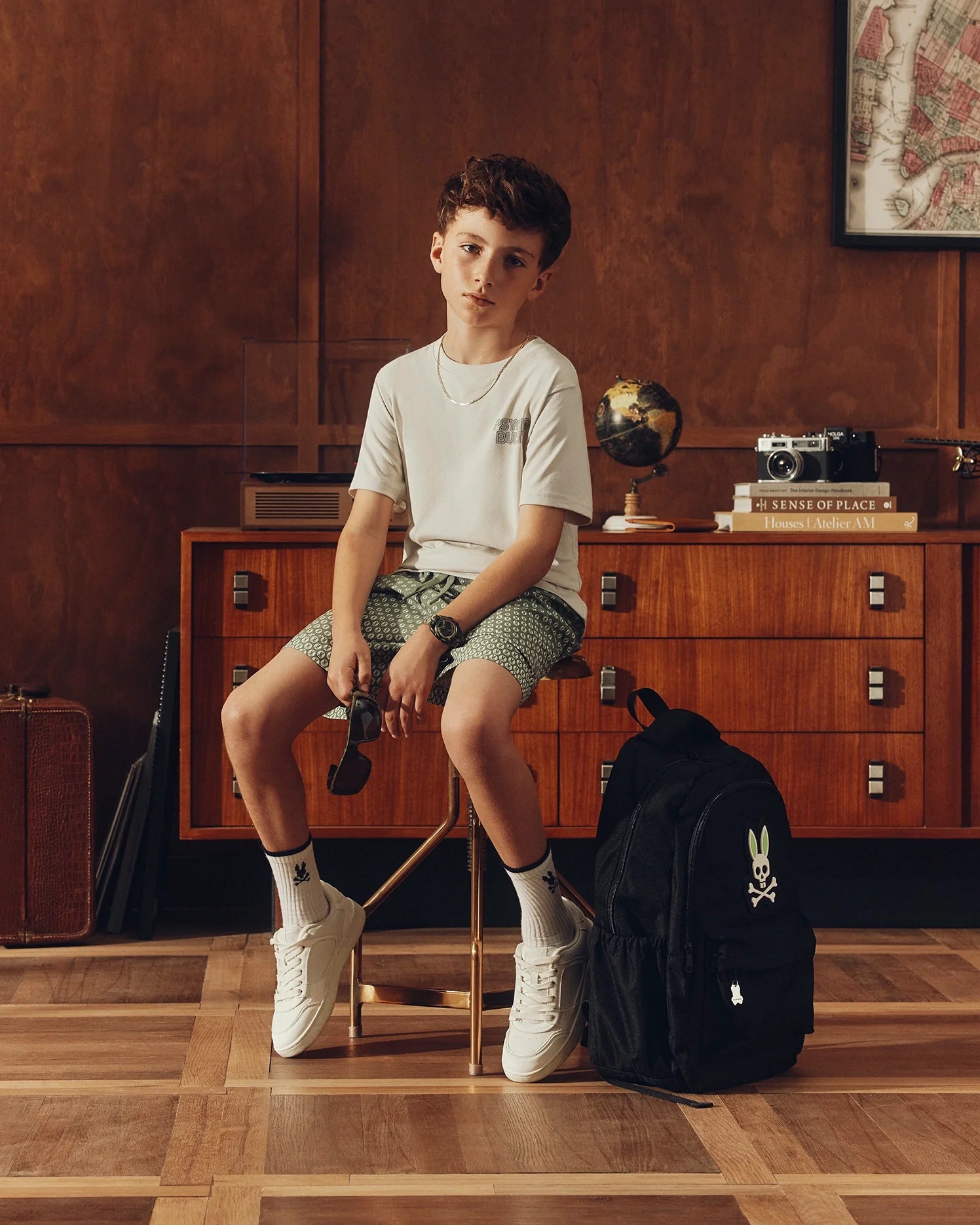 A young boy sits on a stool in a vintage-inspired room, wearing a Psycho Bunny KIDS SYDNEY BACK GRAPHIC TEE in white Pima cotton, green shorts, and white sneakers. A black backpack is nearby. The room includes a wooden cabinet, globe, books, and a camera.
