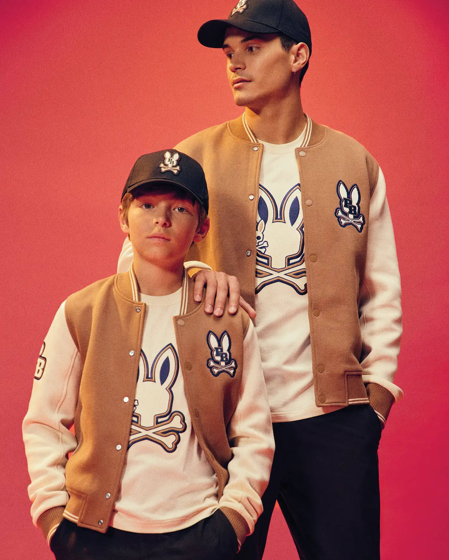 Two people in matching Psycho Bunny KIDS WADE KNIT BOMBER jackets, colorblocked varsity style with bunny skull logos, white logo tees, and black caps pose against a pink backdrop. The taller one rests a hand on the other's shoulder.