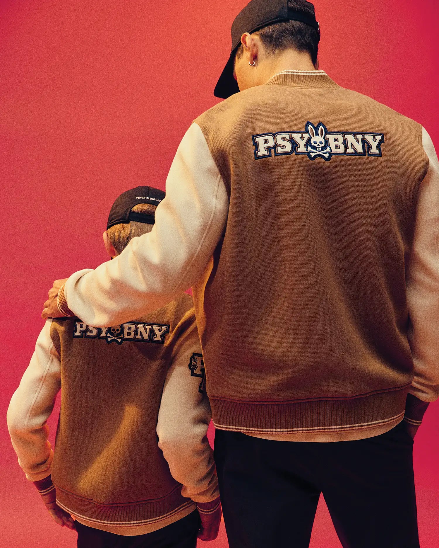 An adult and a child stand side by side, backs to the camera, wearing Psycho Bunny MENS WADE KNIT BOMBER jackets with an embroidered bunny logo and "PSY BN Y" on the back, set against a bold red background.