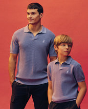 An adult and child stand against a pink backdrop, both in matching light purple Psycho Bunny Leeton Johnny Collar Knit Polos (KIDS LEETON JOHNNY COLLAR KNIT POLO - B0E562F200) with dark pants; the adult smiles as the child looks to the side.