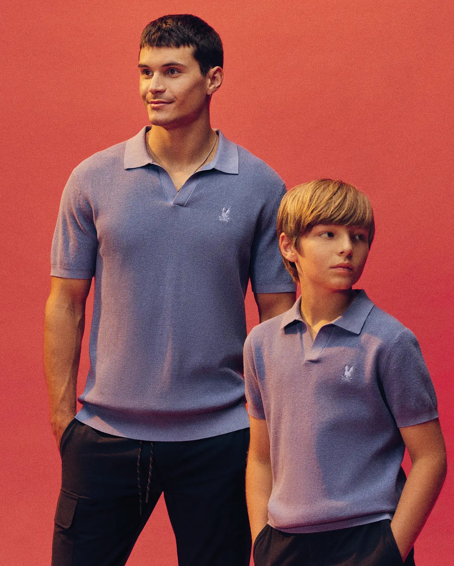 An adult and child stand against a pink backdrop, both in matching light purple Psycho Bunny Leeton Johnny Collar Knit Polos (KIDS LEETON JOHNNY COLLAR KNIT POLO - B0E562F200) with dark pants; the adult smiles as the child looks to the side.