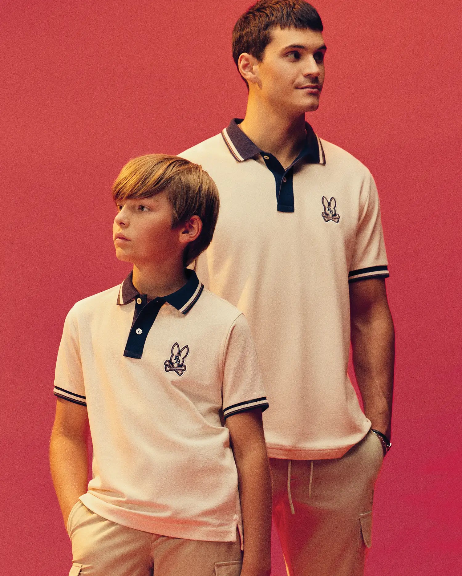 An adult and child wear matching Psycho Bunny MENS WADE PIQUE POLO shirts (MT0100044) in Pima cotton, featuring the embroidered bunny logo, standing against a plain coral background and looking off to the side with relaxed expressions.