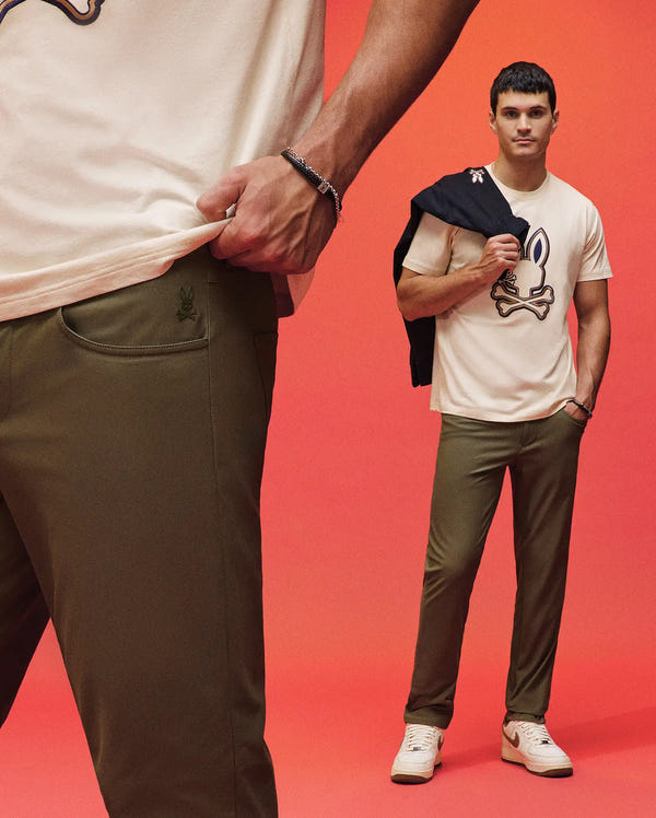 Two men pose against a red background in olive green Psycho Bunny MENS MAVERICK 10K STEP PANTS - B6P788D200 and off-white graphic tees. One holds a jacket; the other shows off the pants’ five-pocket style with a close-up of his outfit and hand.