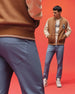 Two men stand against a bright red background wearing brown and beige varsity jackets and Psycho Bunny MENS MAVERICK 10K STEP PANTS (B6P788D200) with five-pocket styling; one faces forward, the other is seen from behind.