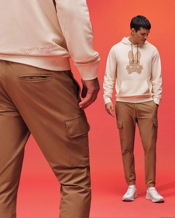 Two men pose against a red background wearing Psycho Bunny MENS LLOYD CARGO JOGGER - B6P159B200 in tan and cream hoodies with an embroidered bunny logo. Both sport white sneakers; one faces forward, the other shows his back.
