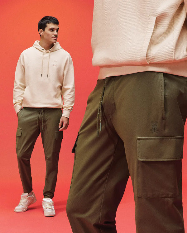 A man stands against a red background in a beige hoodie, white sneakers, and Psycho Bunny’s MENS LLOYD CARGO JOGGER - B6P159B200. The photo spotlights the cargo pants’ embroidered pocket and drawstring in the foreground.