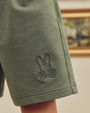 Close-up of Psycho Bunny's olive green Kids Levanzo French Terry Sweatshorts (B0R578F200), made from stretch cotton jersey with an embroidered rabbit head logo above crossed bones, against a background with a blurred framed painting on the wall.