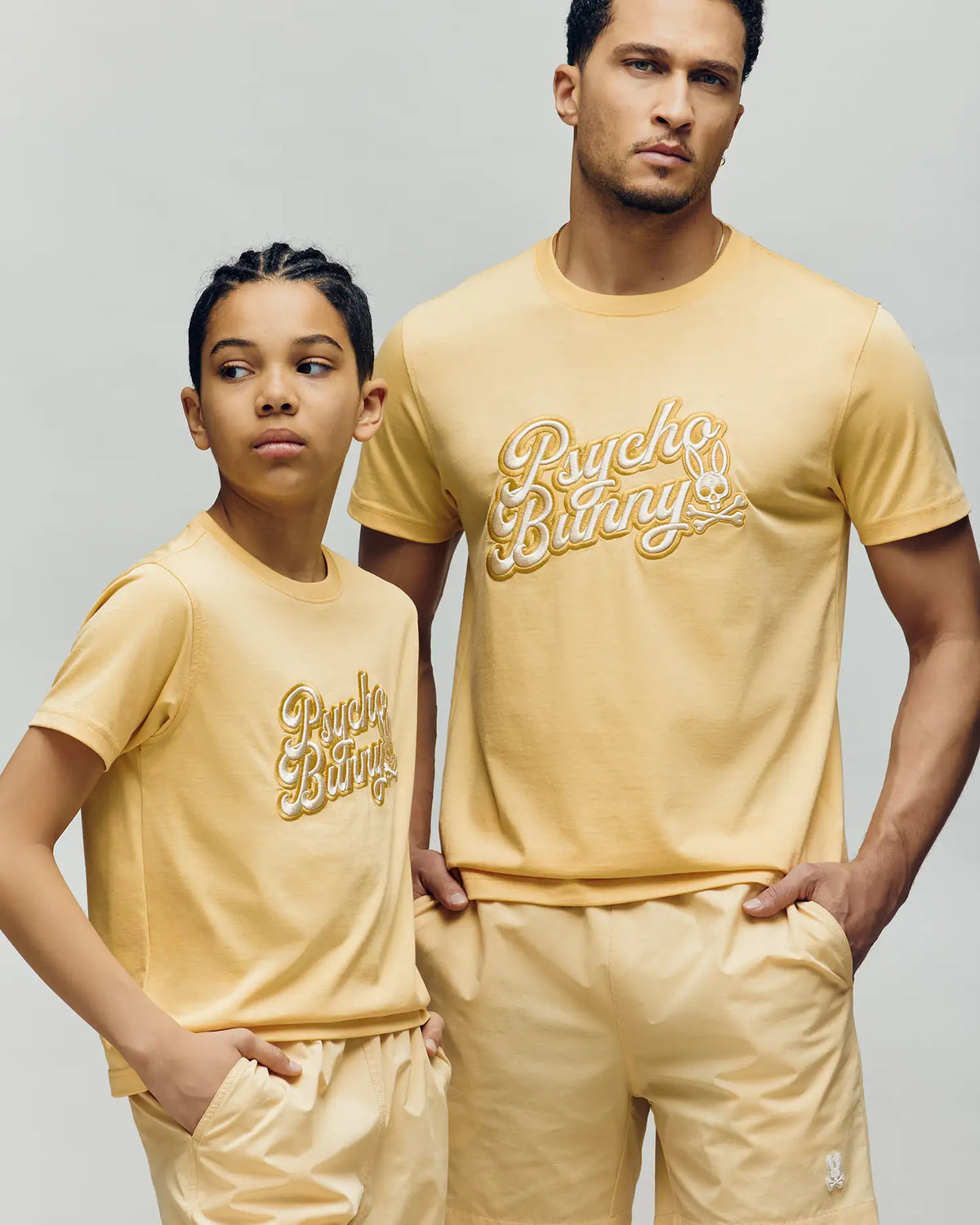 A man and a child wear matching light yellow "Psycho Bunny" MENS ELIAS GRAPHIC TEE (MT0200047) and shorts, both featuring the iconic Bunny graphic, standing against a neutral background while looking in different directions.