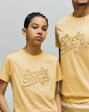 Two people wear matching pale yellow Psycho Bunny KIDS ELIAS GRAPHIC TEEs featuring an embroidered "Psycho Bunny" and Bunny graphic, standing against a plain light background. The younger person stands with one arm bent.