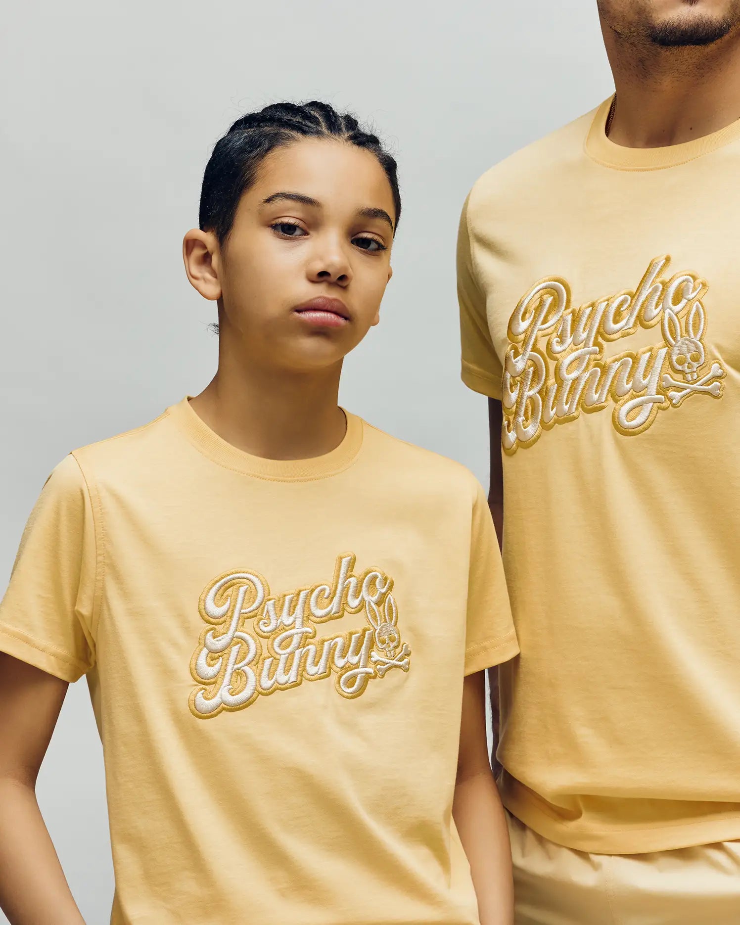 Two people wear matching pale yellow Psycho Bunny KIDS ELIAS GRAPHIC TEEs featuring an embroidered "Psycho Bunny" and Bunny graphic, standing against a plain light background. The younger person stands with one arm bent.
