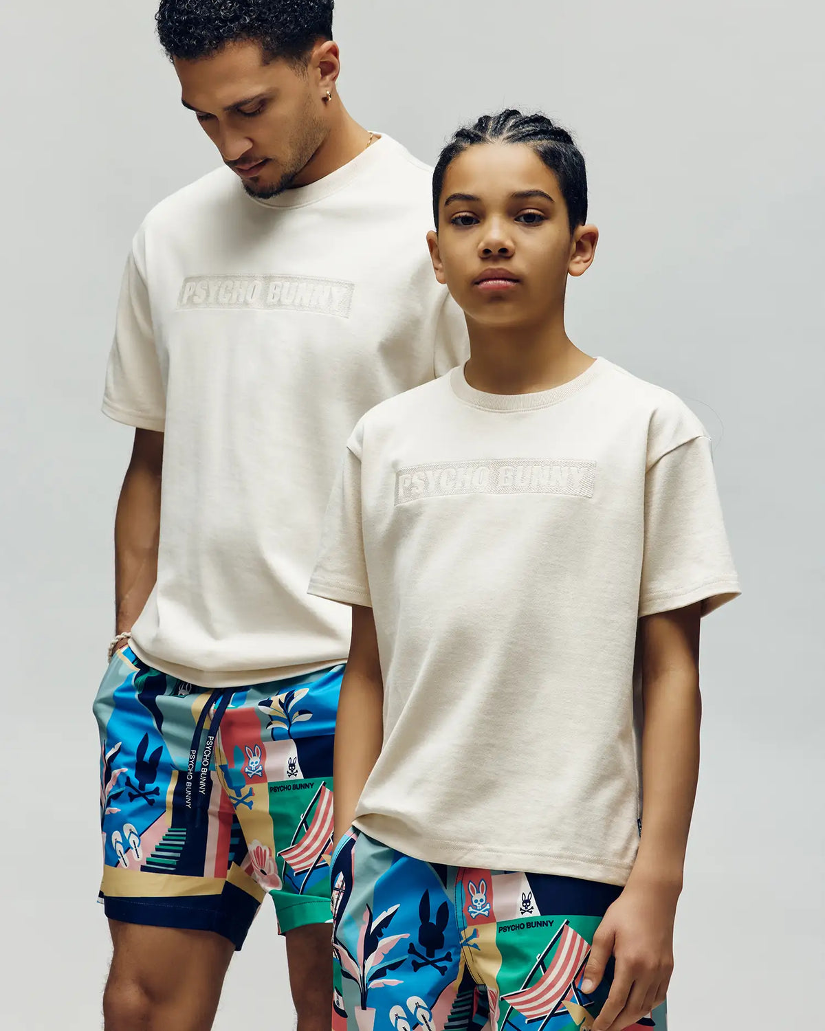 Two kids wear the Psycho Bunny KIDS ZAYN OVERSIZED GRAPHIC TEE (KT0200001) in cream, paired with colorful patterned shorts. Against a plain background, one looks down from behind while the other faces the camera confidently.