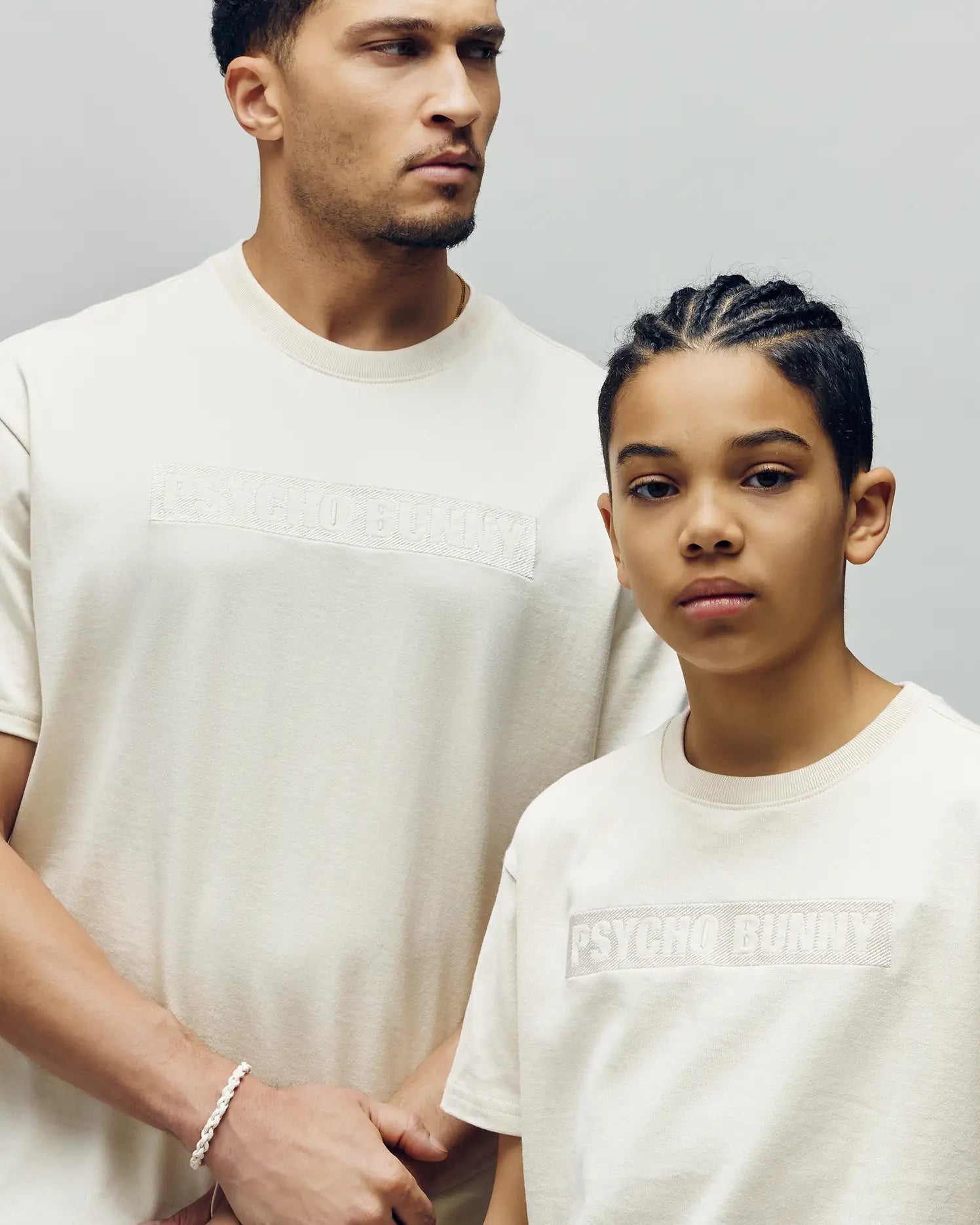 Two people wear off-white Psycho Bunny MENS ZAYN OVERSIZED GRAPHIC TEE - MT0200004 in a relaxed fit; one, taller, looks aside while the shorter person with braids faces the camera, both posing against a plain background.