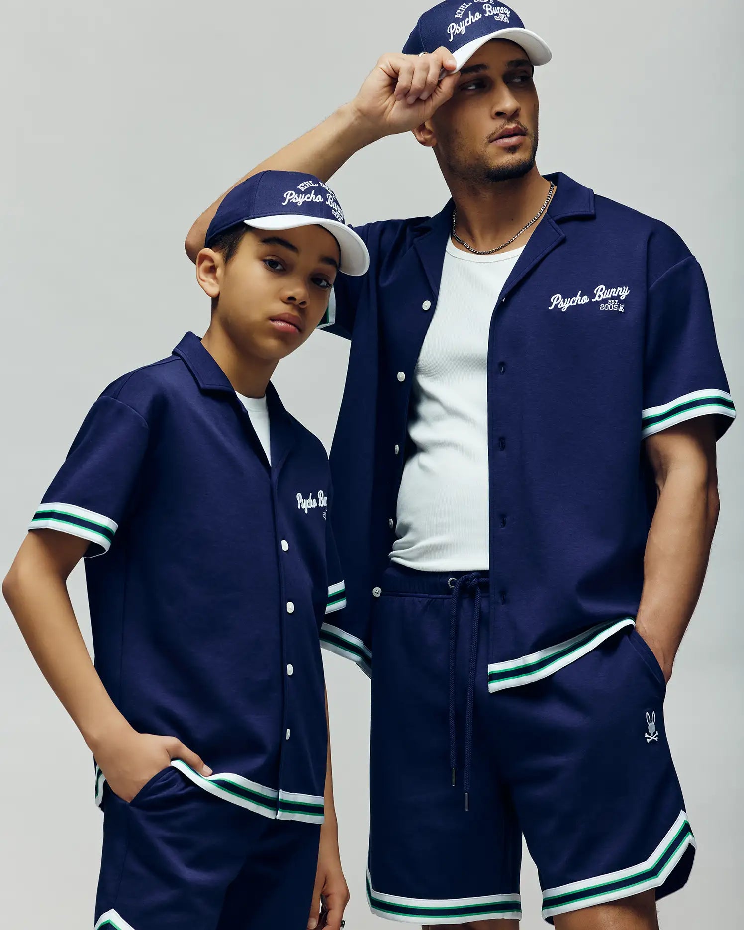 Two people wear matching Psycho Bunny navy KIDS BASIL TAPED CAMP COLLAR shirts and shorts, made from a breathable, flexible premium cotton-modal blend. The adult rests his arm on the boy’s shoulder.