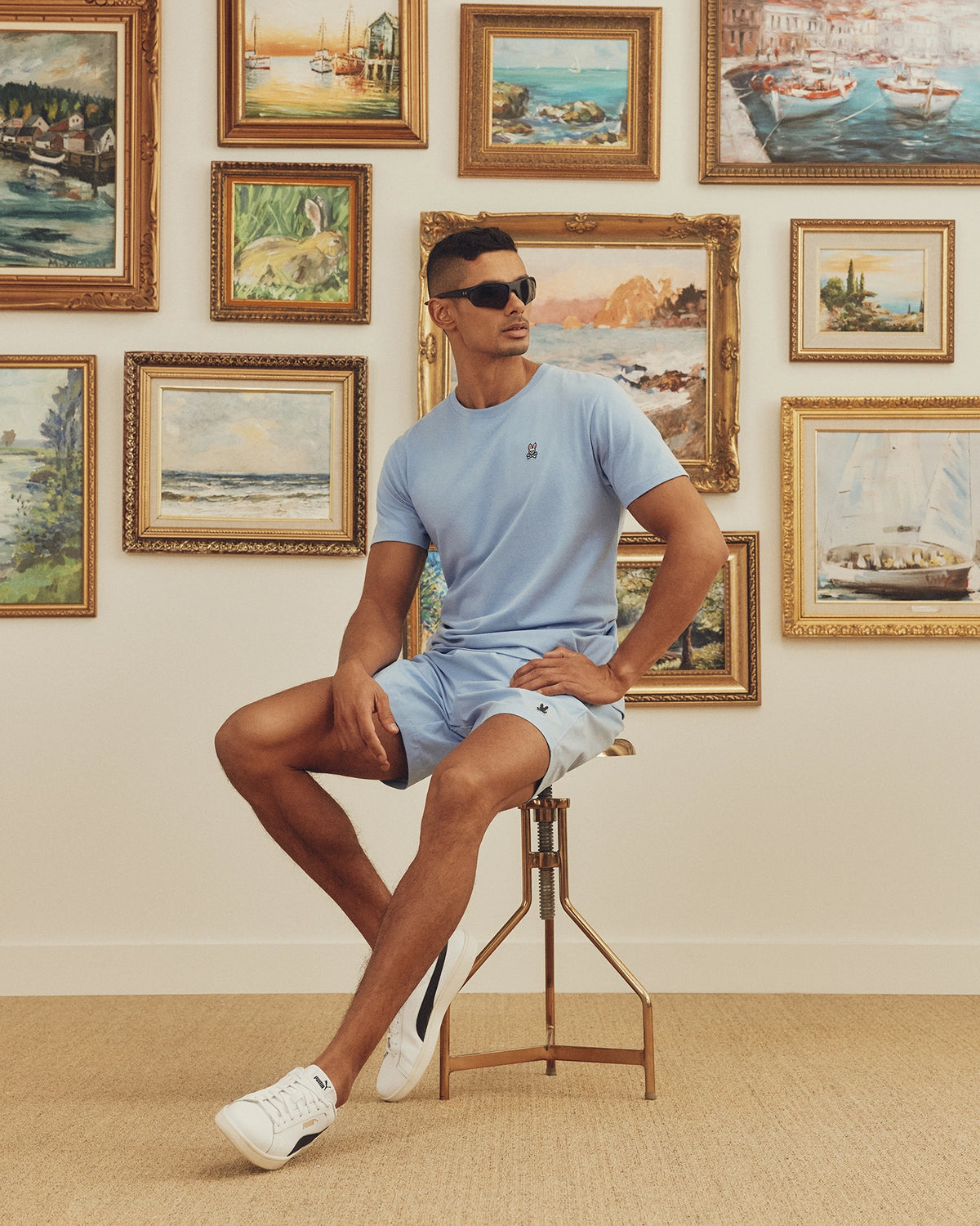 Wearing a Psycho Bunny MENS CLASSIC CREW NECK TEE - B6U014B200 in light blue and sunglasses, a person sits stylishly on a tall stool before a wall of framed landscapes and seascapes, legs crossed with one arm resting on their knee.